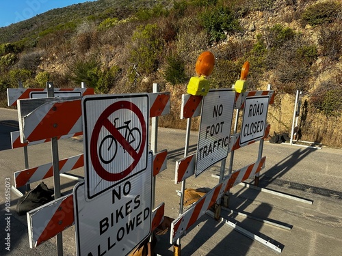 Barriers block all traffic�cars, bikes, and pedestrians�on California Highway 1. Signs mark the closure and prohibit access, set against the backdrop of rugged terrain.