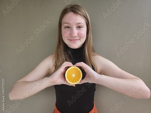 Adolescente loira segurando metade de uma laranja, fazendo um coração com as mãos e a laranja no meio, sorrindo e com fundo neutro. Foto de retrato.