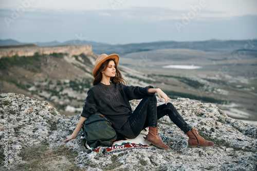 Adventurous woman with backpack and hat sitting on mountain edge enjoying scenic view