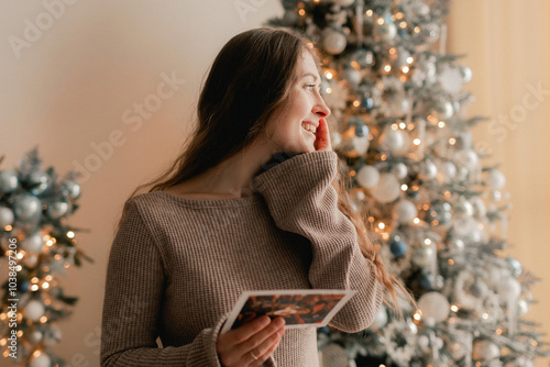 Side view of charming woman reading Christmas greeting card on background of Xmas tree at cozy living room with festive interior. Surprised lady pleasure reading letter at home. Part of a series