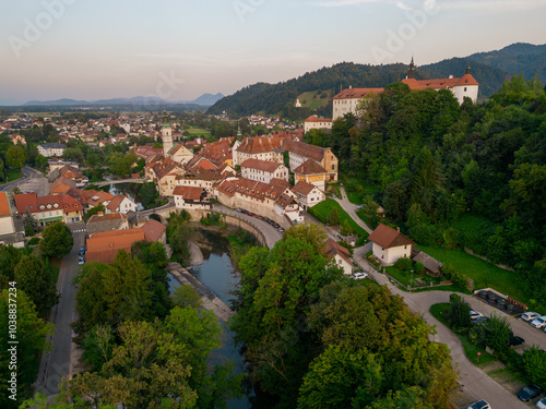 Green riverbank with trees in Skofja Loka, Slovenia. Aerial view above Sora river towards historic town with castle on the hill at sunset. Medieval town settled in the valley.