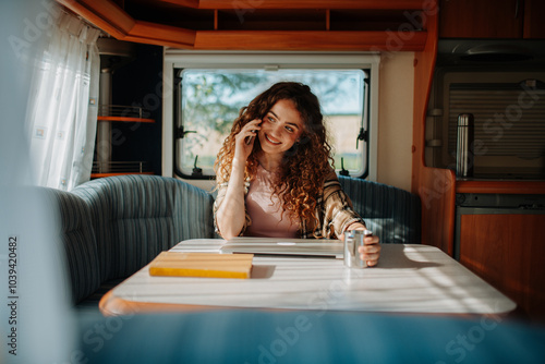 Young beautiful woman is on a camping trip in nature, sitting in her caravan making phone call. Caravan as homeoffice, working remotely.