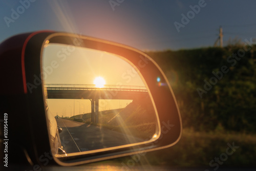 The sun is reflected in the mirror, evening road. View in the side rear view mirror of a red car driving along the highway