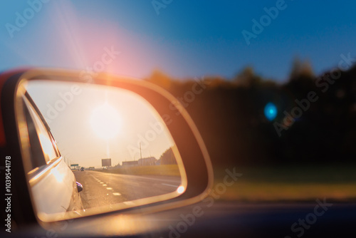 The sun is reflected in the mirror, evening road. View in the side rear view mirror of a red car driving along the highway