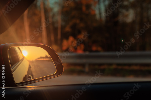 The sun is reflected in the mirror, evening road. View in the side rear view mirror of a red car driving along the highway