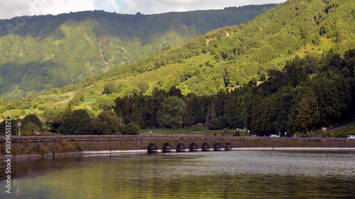 The photo shows a bridge over a huge blue-green lake, surrounded by the green slopes of the Portuguese Azores. The photo is taken from a distance, showing nature and a blue sky.