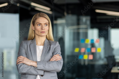 Mature business woman in gray suit standing confidently with arms crossed in modern office. Her composed demeanor conveys leadership, professionalism, and authority in workplace.