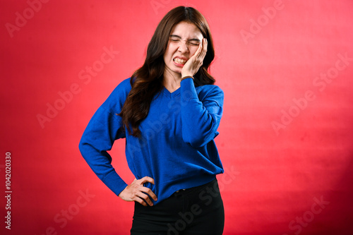 An Asian woman with long brown hair, wearing a blue sweater, is wincing in pain with her hand on her cheek and her other hand on her hip. She stands against a red background, looking frustrated