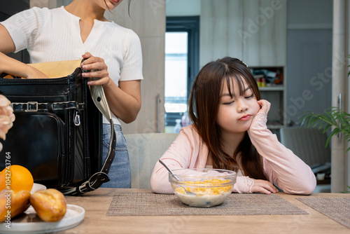 An Asian girl sits at the dining table while her mother packs her school bag, refusing to eat breakfast before going to school.