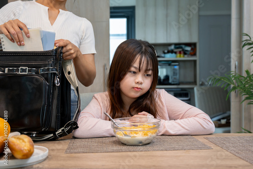 An Asian girl sits at the dining table while her mother packs her school bag, refusing to eat breakfast before going to school.