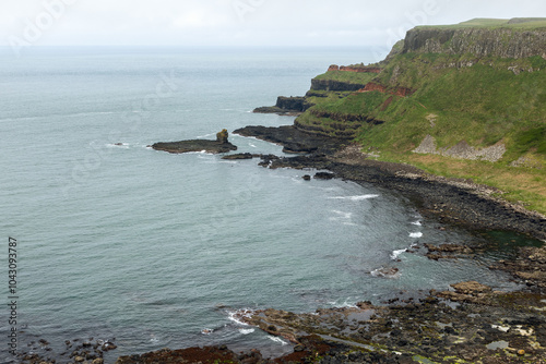 A serene coastal view of Giant's Causeway, showcasing the interplay between green cliffs, rocky shores, and the calm ocean. The layered cliffs add depth to the scene