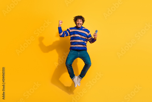 Full length photo of handsome young guy jump hold device winning dressed stylish blue striped garment isolated on yellow color background