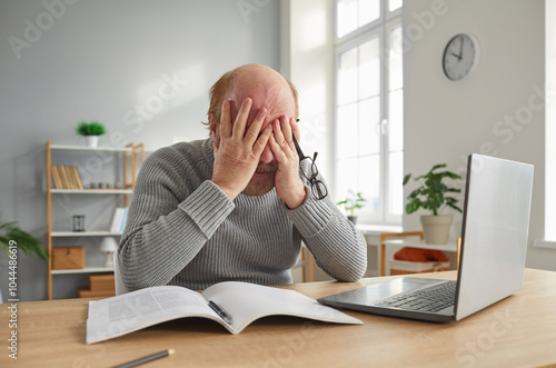 Elderly man working from home office with a laptop and documents, stressed and sad due to a headache. He struggles to focus on tasks while enduring pain and discomfort in a quiet work environment.