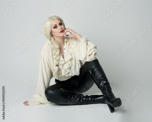 full length portrait of beautiful female model, wearing vintage gothic fantasy ruffle blouse white shirt, leather pants, blonde hair holding knife weapon. sitting pose, isolated on studio background.