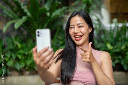 A smiling Asian woman is enjoying a video call on her smartphone while sitting at an outdoor table.