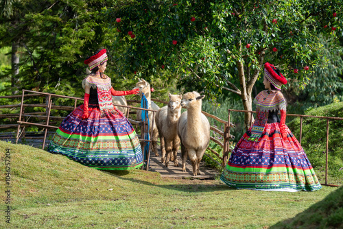 Two women in colorful traditional dresses interact with alpacas in a rural outdoor setting, surrounded by greenery and trees.