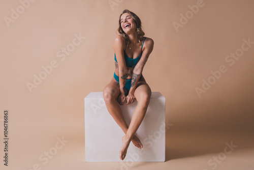 Full body photo of attractive young girl sit white cube laughing wear cyan fashion lingerie isolated beige background no filter