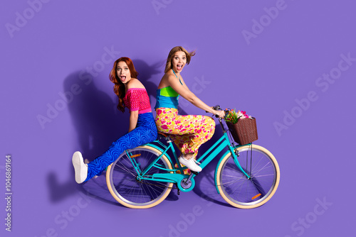 Full body portrait of two pretty impressed young girls ride bike empty space ad isolated on purple color background