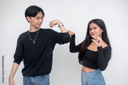 A young asian woman mocks the small size of a man's arm while touching it, gesturing it to be scrawny and small. Isolated on a white background.