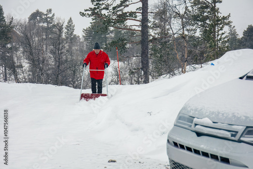 Man in red jacket pushing a snow pusher on a snow-covered road by mountain cabin ,winter task, outdoor maintenance, rural winter