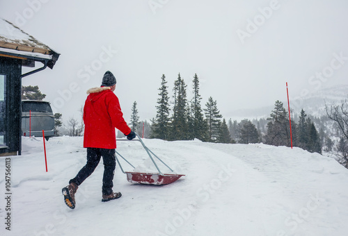 Man in red jacket pushing a snow pusher on a snow-covered road by mountain cabin ,outdoor task, snow removal, rural lifestyle, winter work