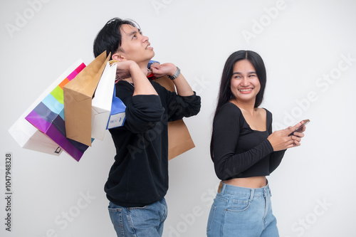 A young man is annoyed and overburdened by carrying a lot of shopping bags for his spendthrift girlfriend, who is on her cellphone. Isolated on a white background.