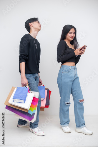 A young man is annoyed and overburdened by carrying a lot of shopping bags for his spendthrift girlfriend, who is on her cellphone. Isolated on a white background.