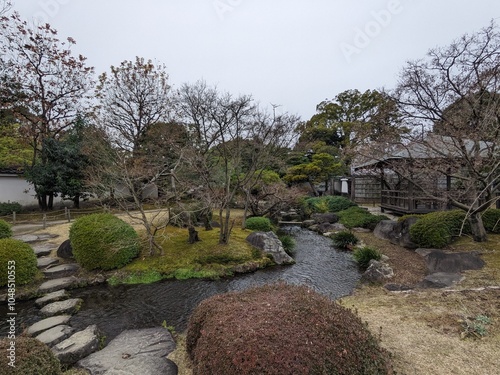 Himeji, Japan - 12.19.2023: A stone pathway over a stream next to a wooden pavilion in a traditional Japanese garden in Koko-en surrounded by manicured trees under a cloudy sky