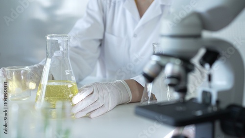 A scientist, wearing a lab coat and white protective gloves, is pouring a yellow oily liquid from one beaker to another near microscope in laboratory, close up. Medicine and science concept