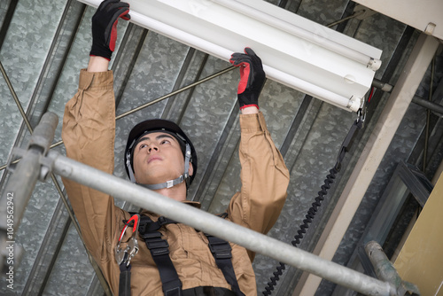 足場を使って蛍光灯を変える安全帯をつけた作業着の人 電気工事や電気工事士のイメージ