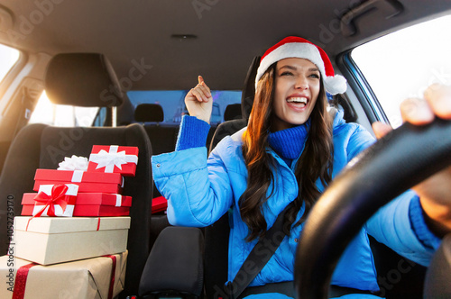 Overjoyed young woman in Santa Hat driving car with Xmas gift boxes nearby and enjoy music, singing song feeling enjoyment of winter holiday preparation