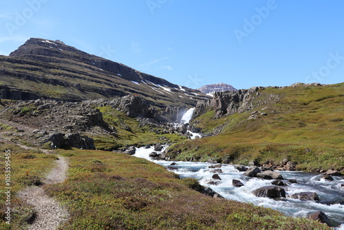 unspoiled landscape with a narrow hiking trail next to a wild mountain river with a waterfall in the distance