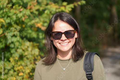 Young smiling girl in khaki t-shirt standing in forest with backpack over shoulder. Concept of unity with nature in travel