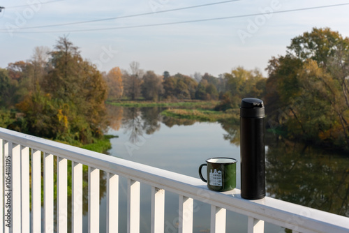 Thermos and mug of tea on railing overlooking picturesque river on sunny fall day. Concept of outdoor adventure, walking and enjoying nature with warm drink. High quality photo