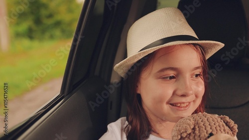 Happy female child face in summer hat laughing smiling adventure automobile enjoy weekend travel vacation. Road trip car travel overjoyed girl kid with bear toy riding open window closeup slowmo.