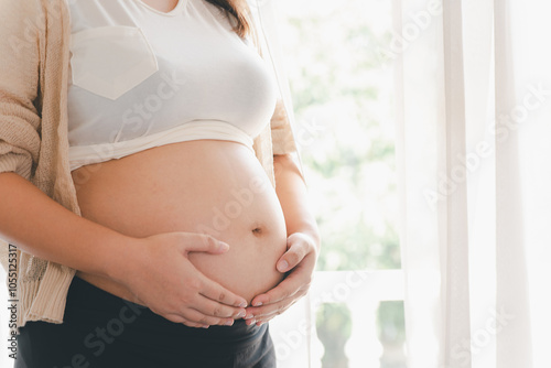 Close-up of a pregnant woman gently holding her bare belly, standing by a sunlit window, symbolizing motherhood and anticipation.