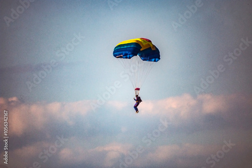 A parachutist flies on a blue and yellow parachute in a cloudy sky