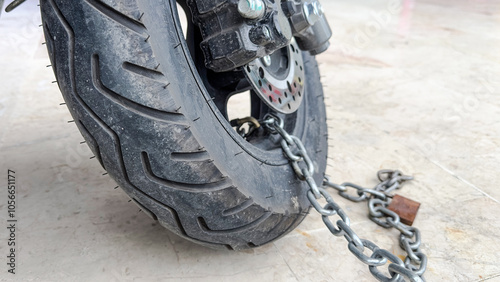 Close-up of a motorcycle wheel secured with a heavy-duty chain and lock, indicating security and anti-theft measures on a concrete surface.