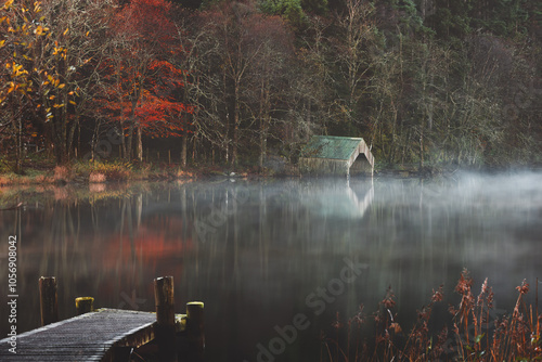 A misty autumn morning at Loch Ard with a serene boathouse and reflections, surrounded by fall foliage in the Scottish Highlands.