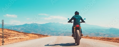 Motorcyclist riding on an empty road with mountains in the background under a clear blue sky.