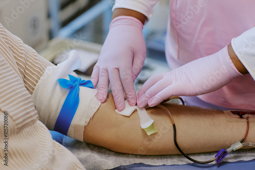 Medical professional wearing pink gloves taking blood sample from patient's arm with blue tourniquet applied. Sample collection process happening in clinical environment