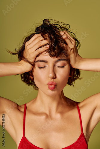 A young woman holds her head, showing a mix of emotions while playfully pouting her lips.