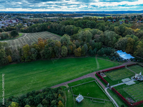 The scene captures an expansive autumn landscape featuring lush greenery intermixed with trees showcasing fall colors. Fields stretch across the terrain, while a nearby gathering area is visible.