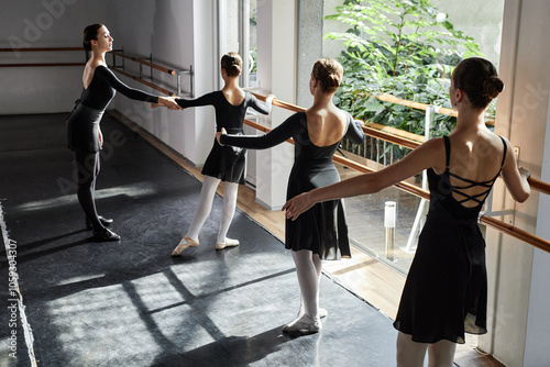 Rear view of three young girls wearing elegant dance leotards learning ballet positions at barre while female choreographer assisting children in sunlit studio