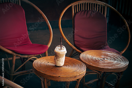 Iced coffee in a single use plastic container set among rattan table and chair showing the irony between eco-friendly furniture and plastic polluting container