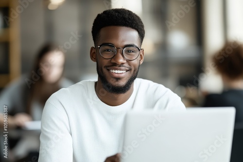 A young man with glasses is smiling while using a laptop in a lively cafe, representing a blend of modern work, sociability, and casual ambiance.