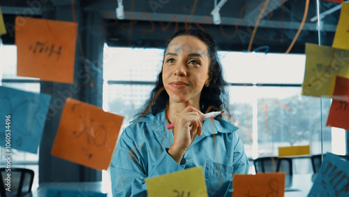 Portrait of skilled young beautiful hispanic project manager thinking about marketing idea. Professional businesswoman making business plan by using sticky notes at glass board in office. Manipulator.