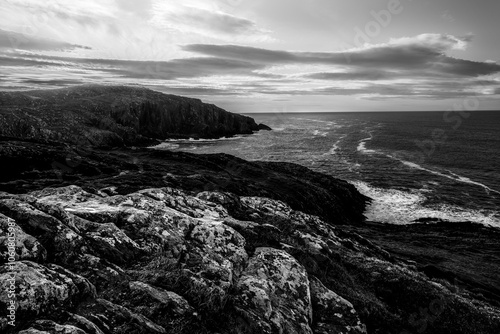 This striking black and white capture showcases the rugged coastline, where rocky cliffs plunge into the restless sea, all set against a moody sky just after sunset.