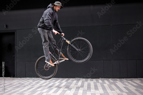 young hipster guy doing stunts on a bike in the city against the background of a black wall, street rider shows freestyle on a bike and makes dangerous moves