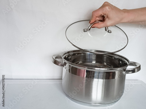 Female hand of housewife opens glass lid of stainless steel saucepan. Kitchen metal utensils stands on table. Background, place for text.